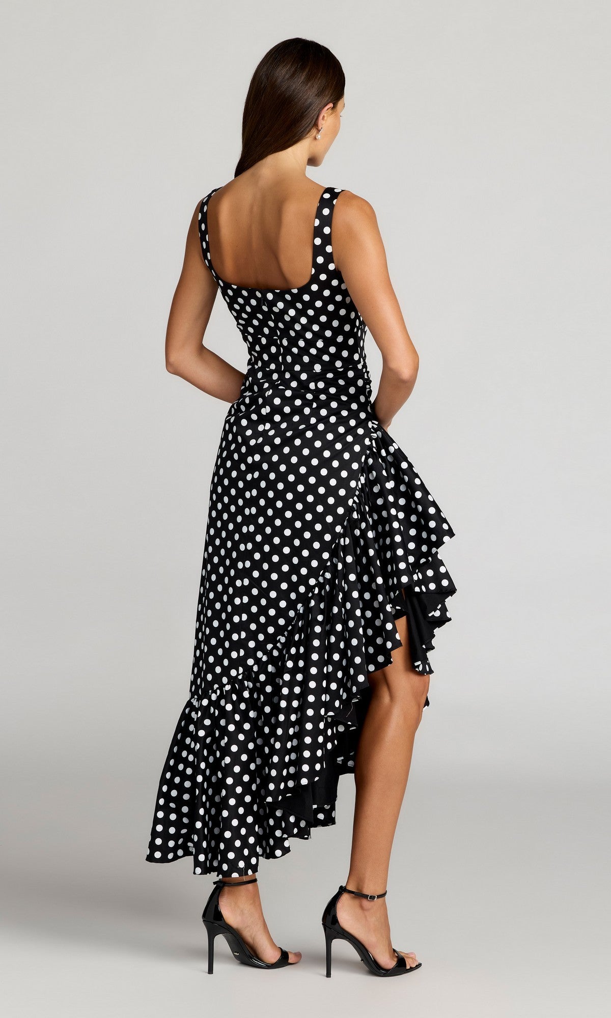 A woman stands with her back turned, wearing the Mac Duggal 27780 midi party dress—a black polka-dot cocktail dress with an asymmetrical ruffled hem—paired with black high-heeled sandals against a plain background.